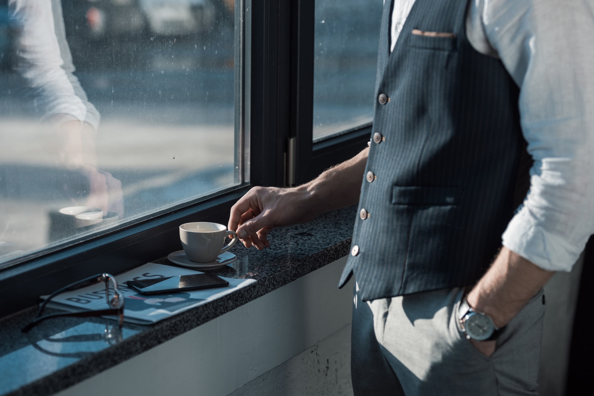 A businessman standing by a window with a coffee cup, tablet, and magazine on the sill, possibly featuring commercial smart window tints like ElectraTint for privacy and glare reduction.